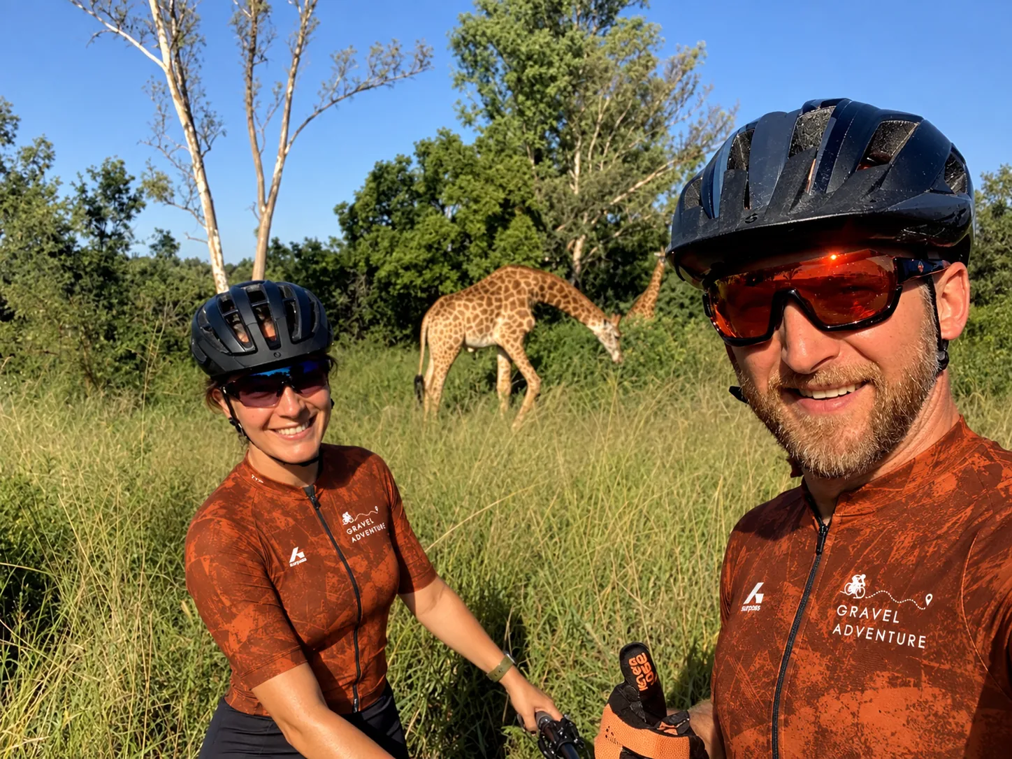Tanya og Michael Sommer, grunnleggerne av Gravel Adventure, på en gravelreise i Sør-Afrika med sjiraffer som beiter i bakgrunnen