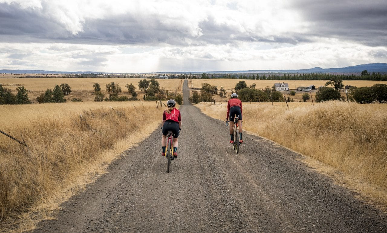 To gravelcyklister kører på en grusvej gennem gyldne marker under en overskyet himmel