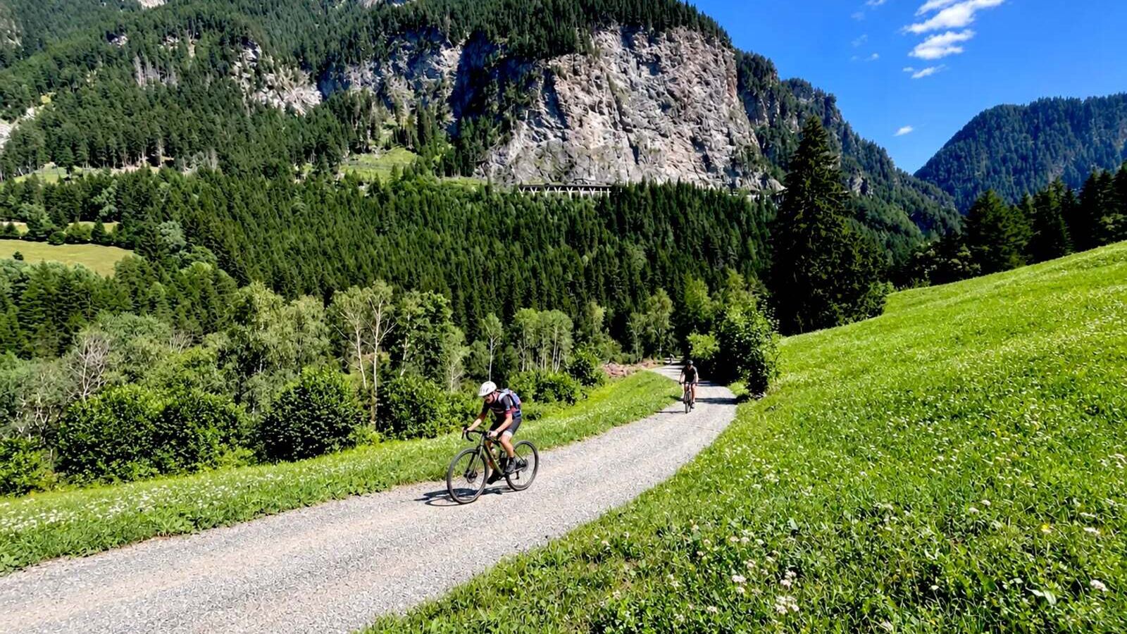 To gravelcyklister kører ned ad en grusvej i en dal mellem skovklædte alpeskråninger på Trans Alp-ruten